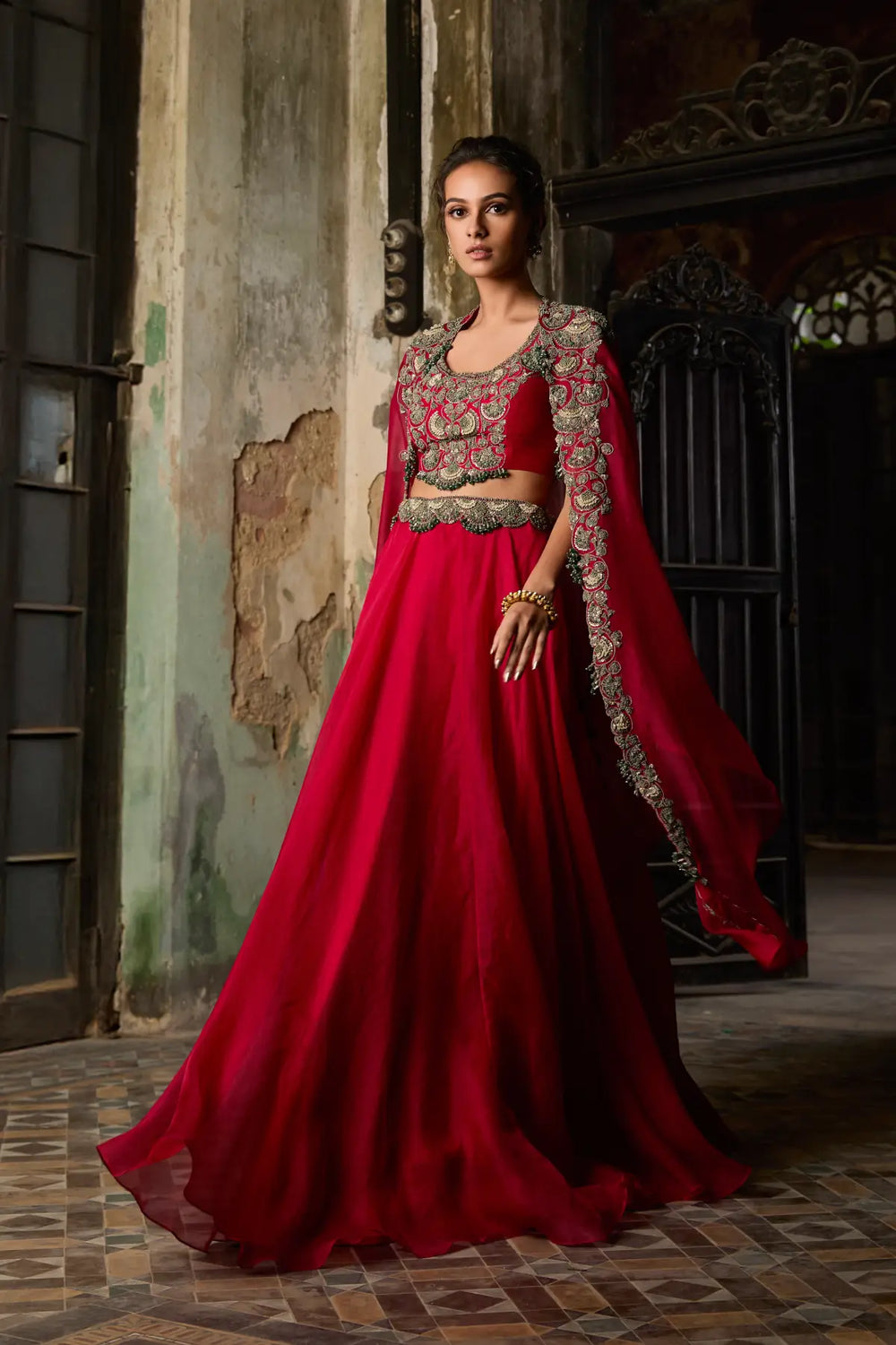 Woman in a red traditional cape and skirt with intricate designs standing in a dimly lit room.