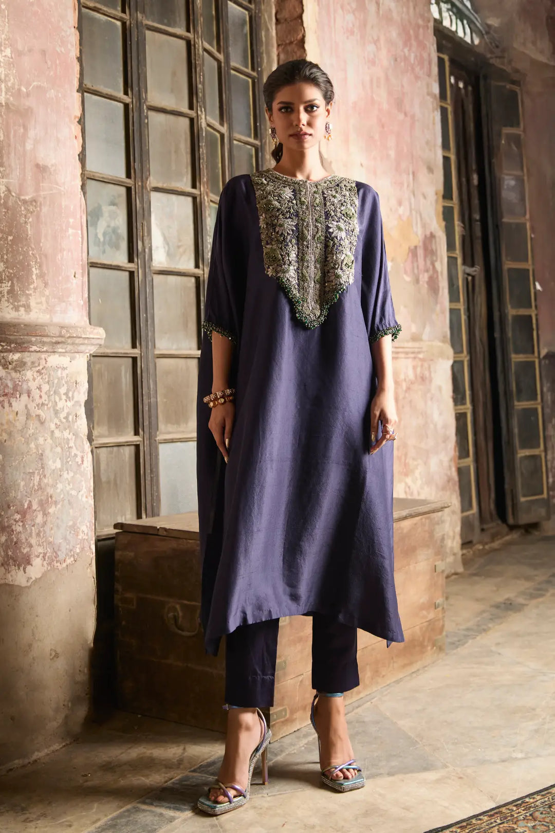 Woman wearing a long, dark blue embroidered kaftan and black pants in an indoor setting.