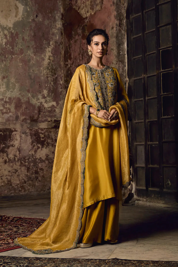 Woman in a traditional yellow sharara with intricate patterns standing against a textured wall.