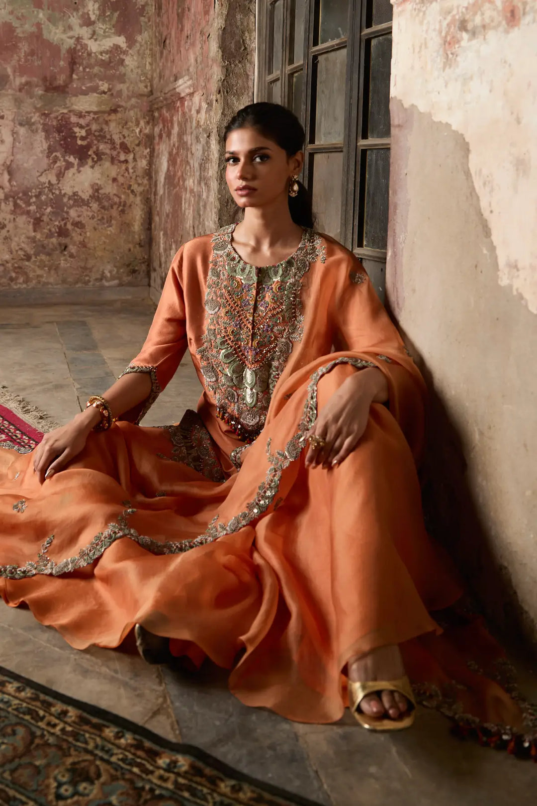 Woman in an orange traditional sharara sitting against a textured wall.