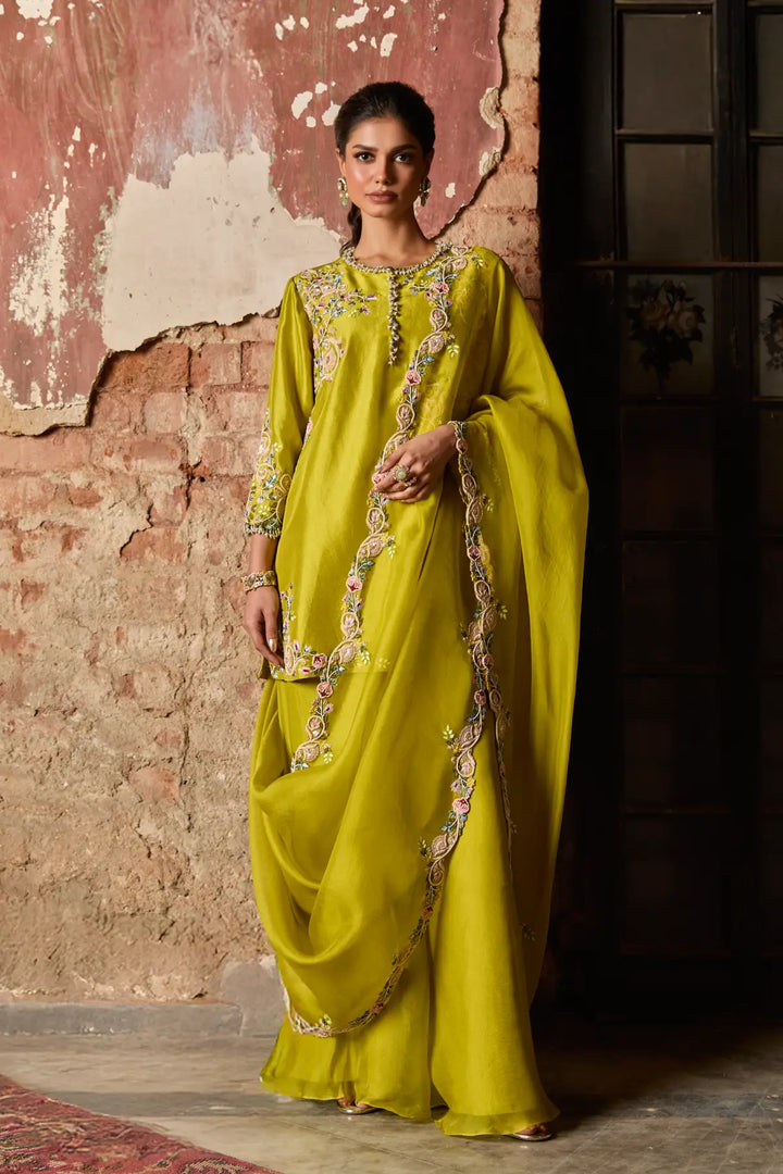 Woman wearing a bright yellow sharara with intricate patterns against a textured wall.