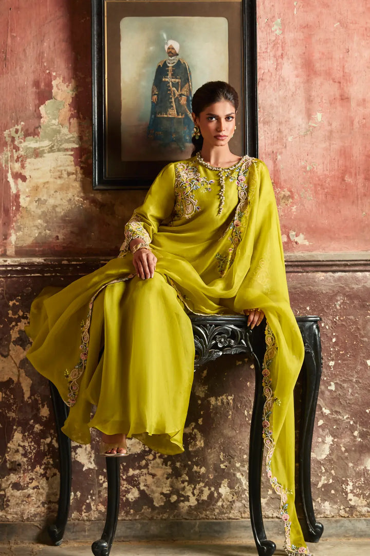 Woman in a yellow traditional sharara sitting on a chair with a textured wall and framed picture in the background.