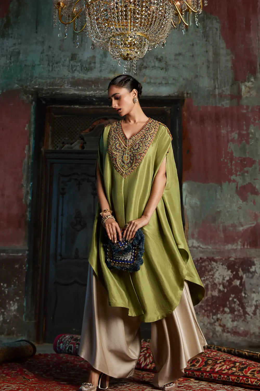 Woman in a green kaftan with intricate detailing standing in a room with a chandelier.