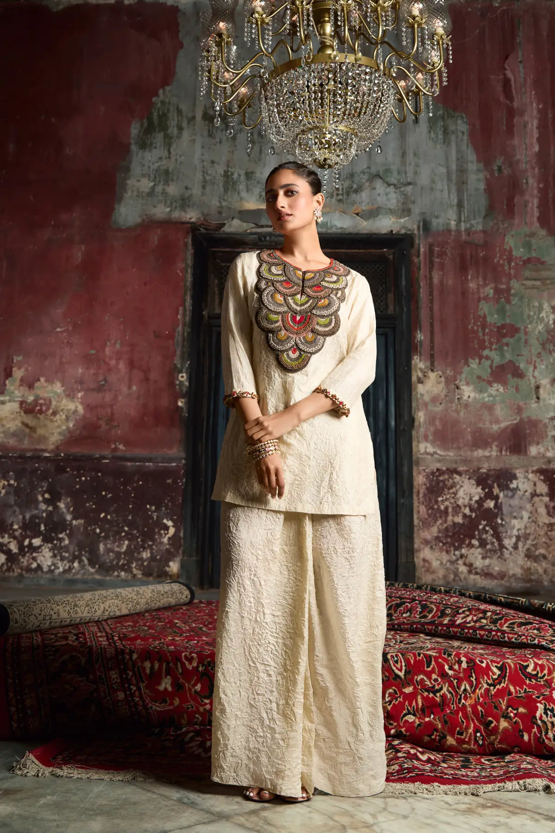 Woman in traditional co-ord set standing in a room with ornate walls and a chandelier.