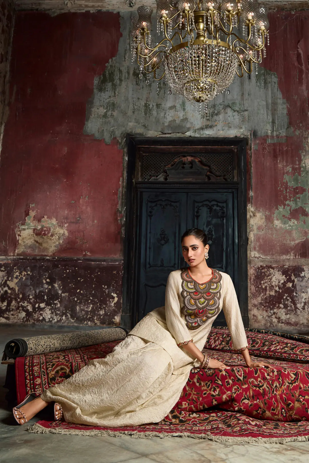 Woman in traditional co-ord set sitting on a patterned rug with a chandelier above.