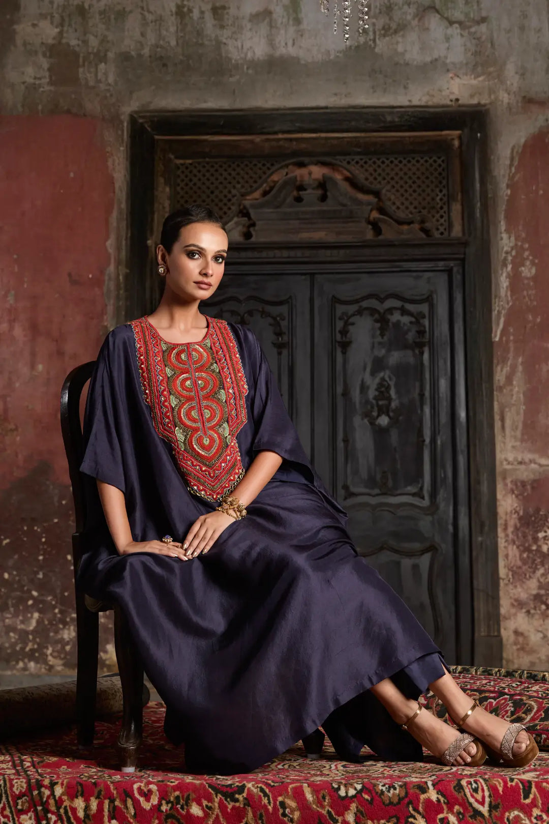 Woman in a dark blue kaftan with red embroidery sitting on a patterned rug.