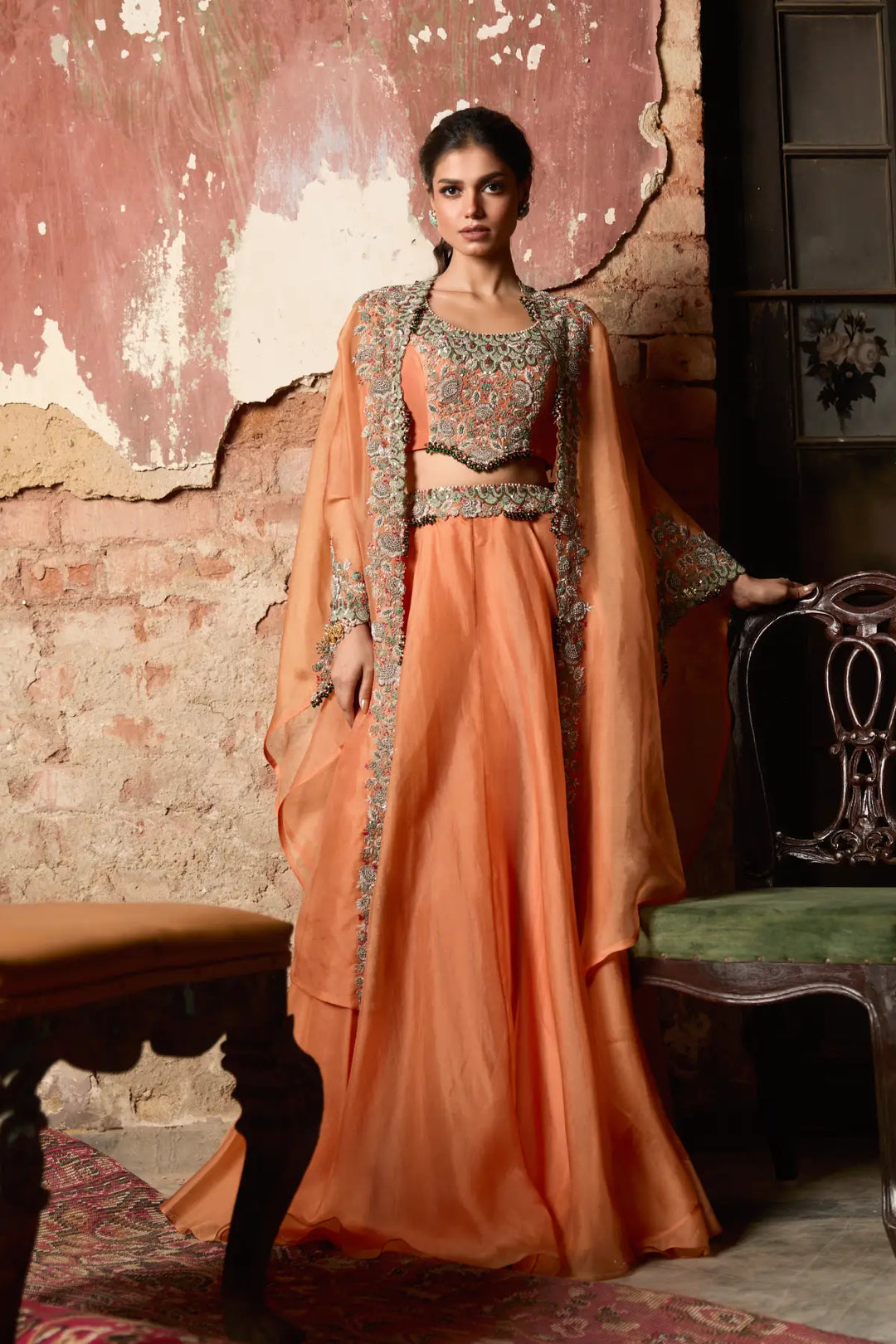 Woman in a peach traditional cape and skirt with intricate designs standing against a textured wall.