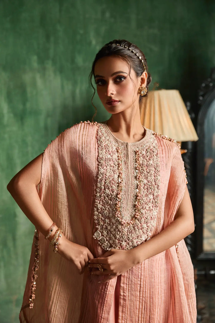 Woman in a peach traditional kaftan with intricate detailing against a green wall.