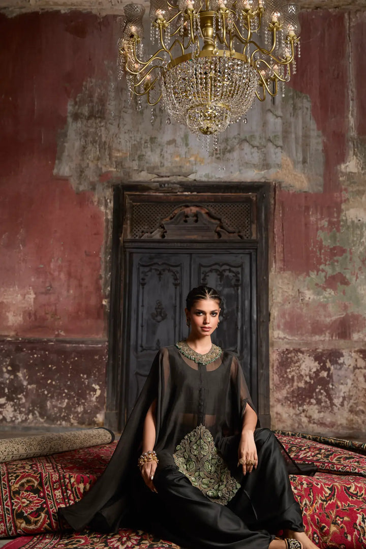 Woman in a black kaftaan sitting on a patterned rug with a chandelier above in an ornate room.