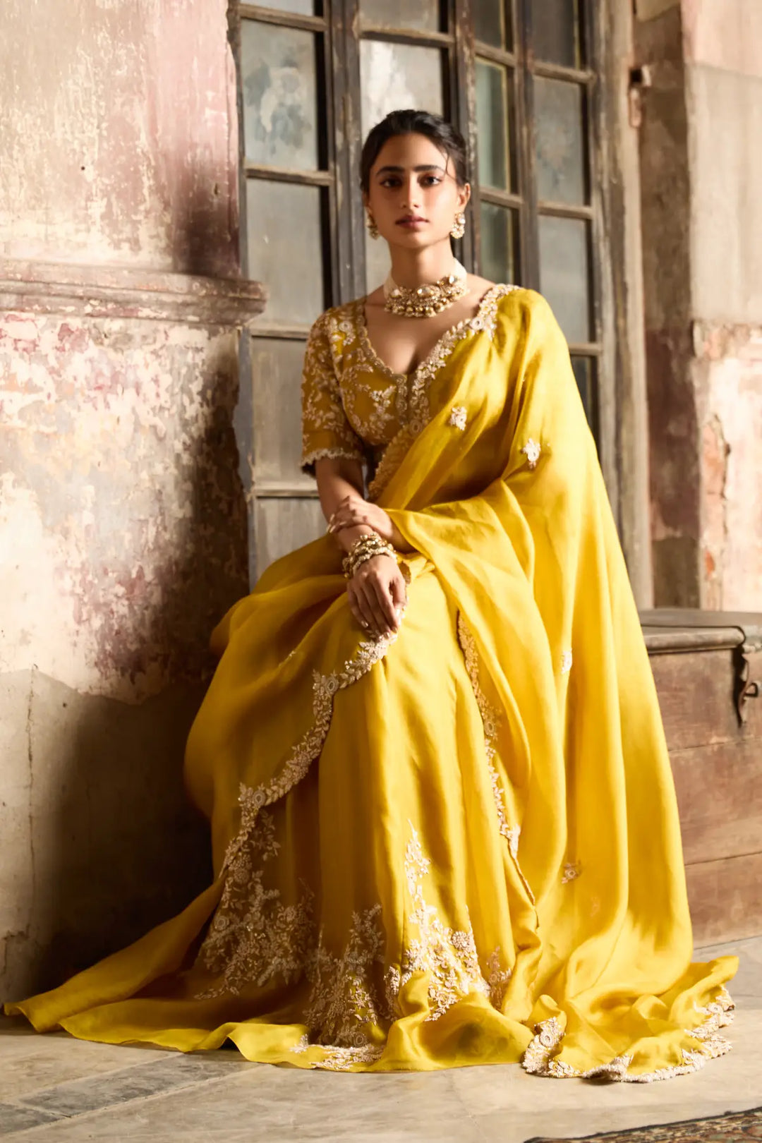 Woman in a yellow saree with intricate designs sitting against a textured wall.