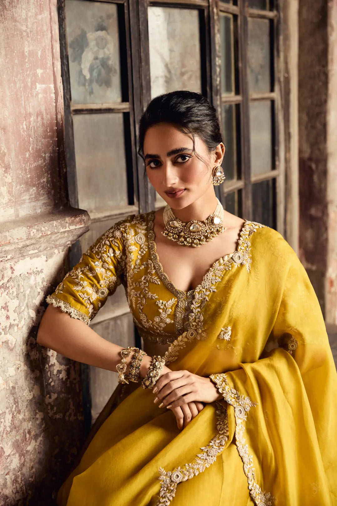 Woman in a yellow traditional saree with gold embroidery sitting against a textured wall.