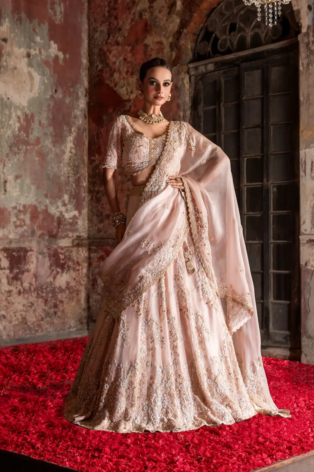 Woman in a traditional lehenga standing on a red carpet with an ornate wall and door in the background.