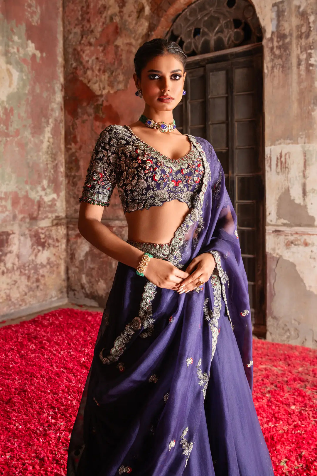 Woman in a traditional lehenga standing in front of a textured wall with red carpet.