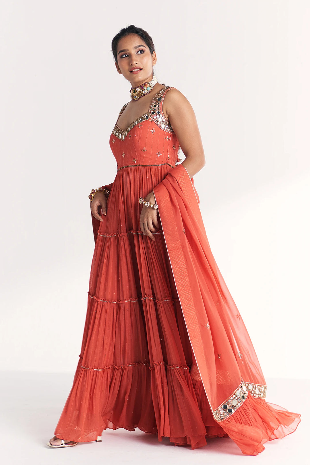 Woman wearing a coral orange traditional Anarkali with intricate detailing on a white background