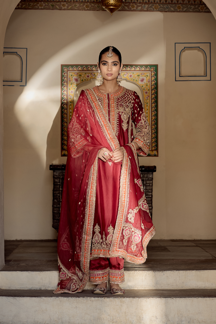 Woman in a traditional red and gold embroidered outfit standing in a decorated room.