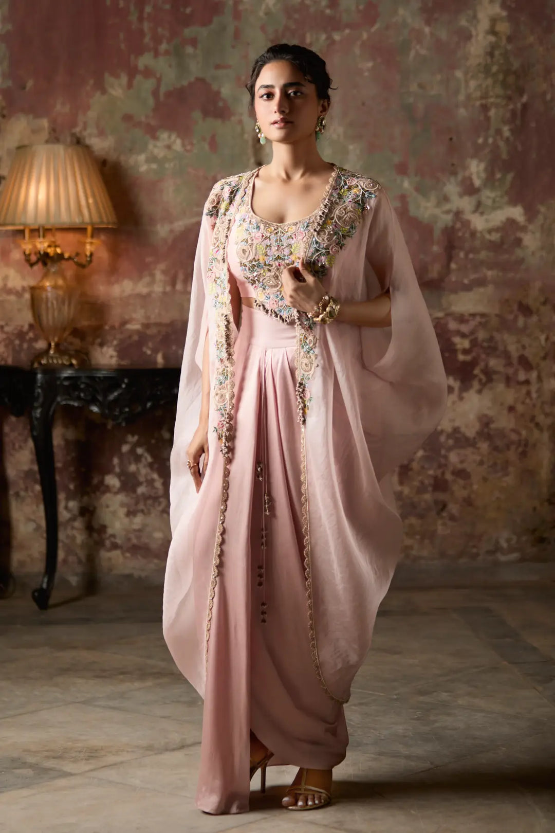 Woman in a pink traditional Skirt Set with intricate designs standing in a room with a vintage lamp.