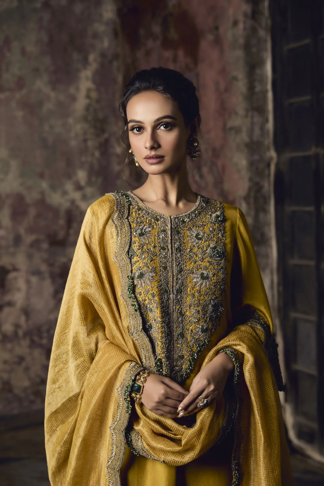 Woman wearing a traditional yellow and gold embroidered sharara against a textured wall.