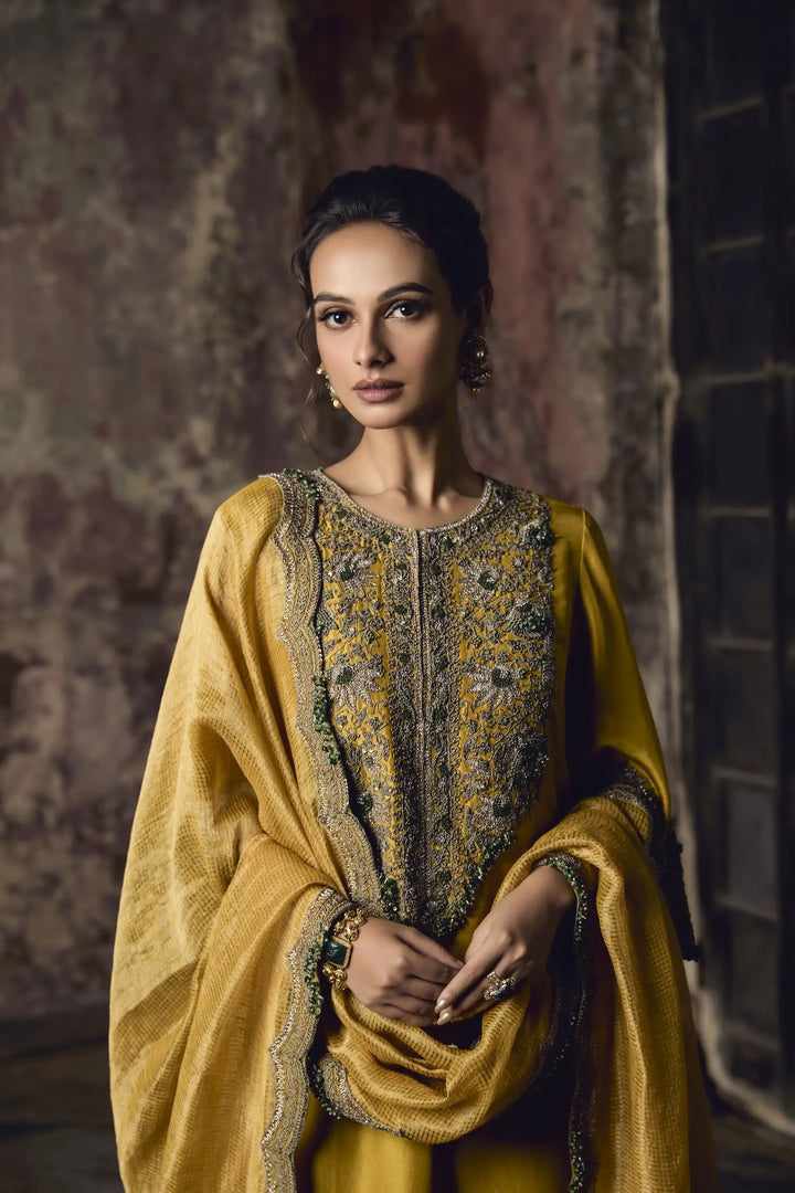 Woman wearing a traditional yellow and gold embroidered sharara against a textured wall.