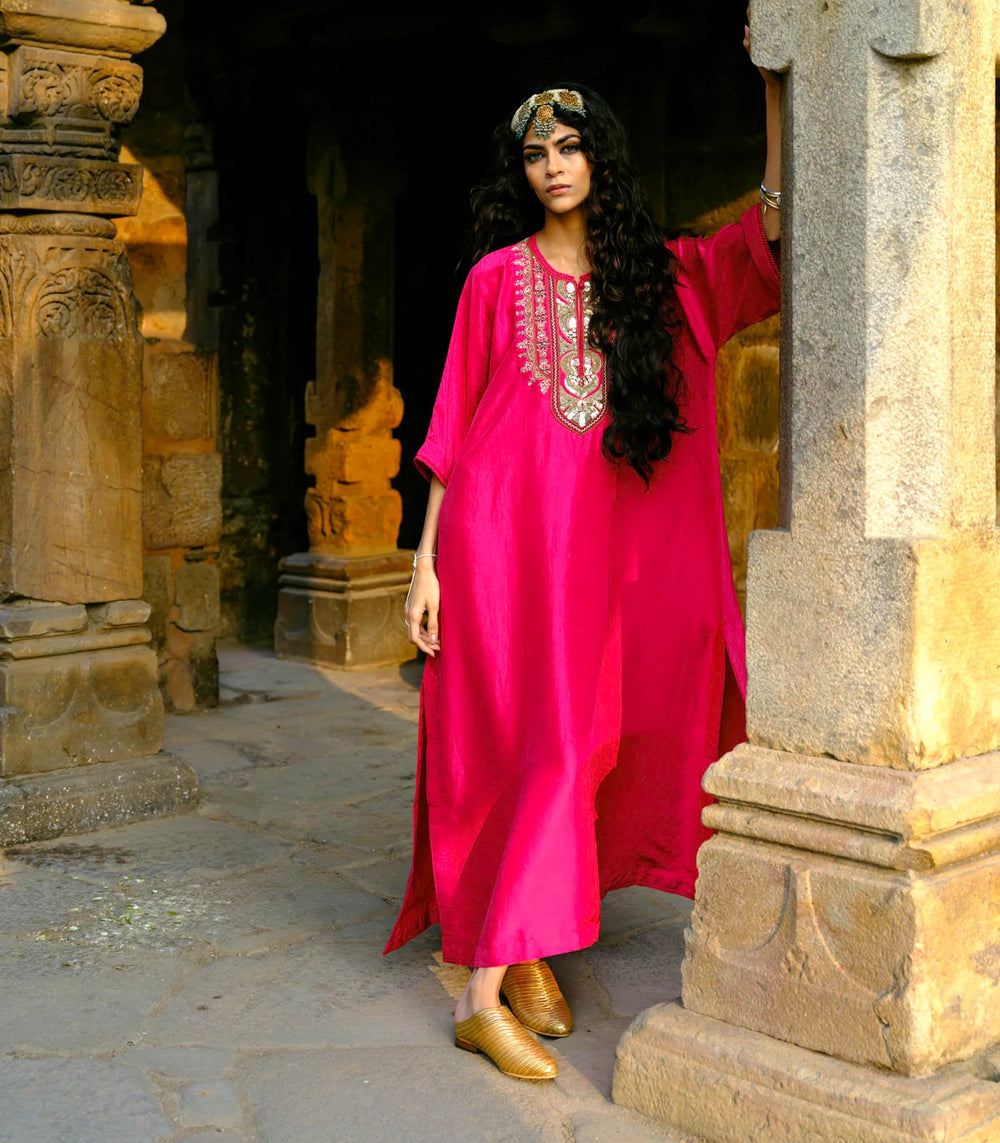 Woman in a bright pink dress with intricate embroidery standing in an ancient stone doorway.