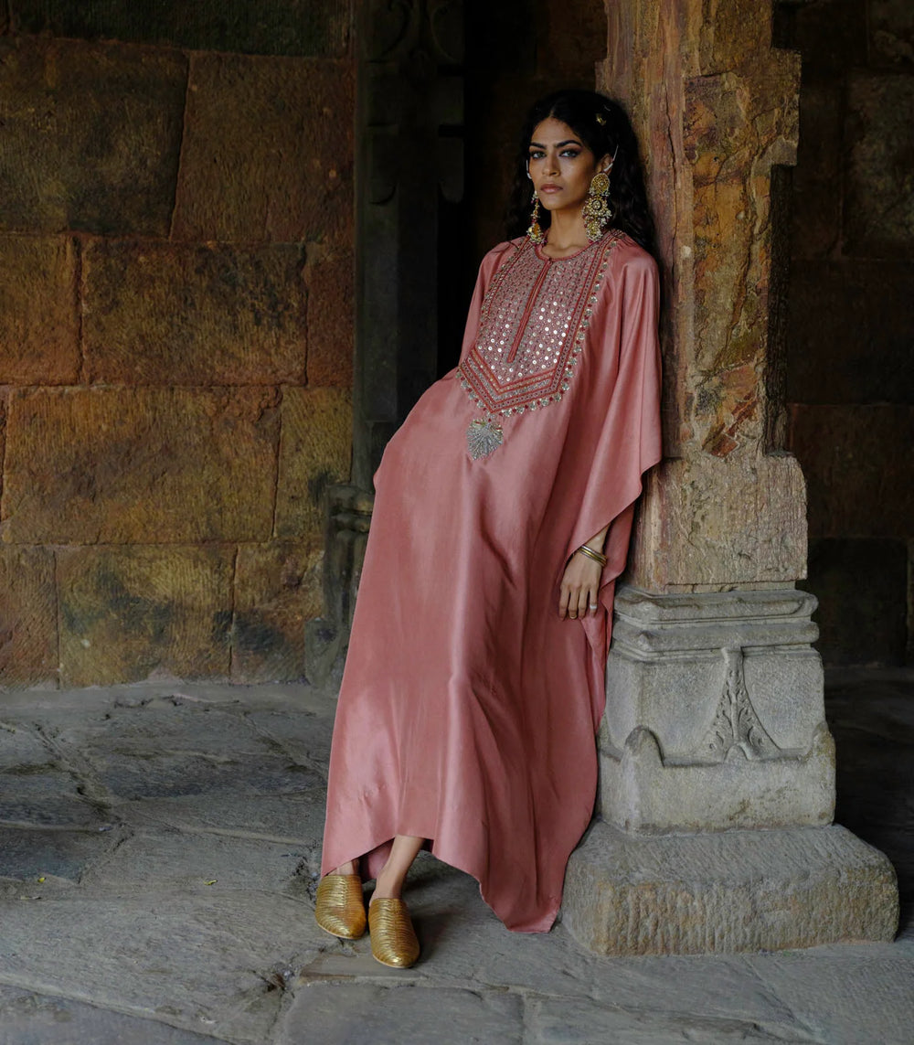 Woman in a pink dress with intricate patterns standing against a stone wall.