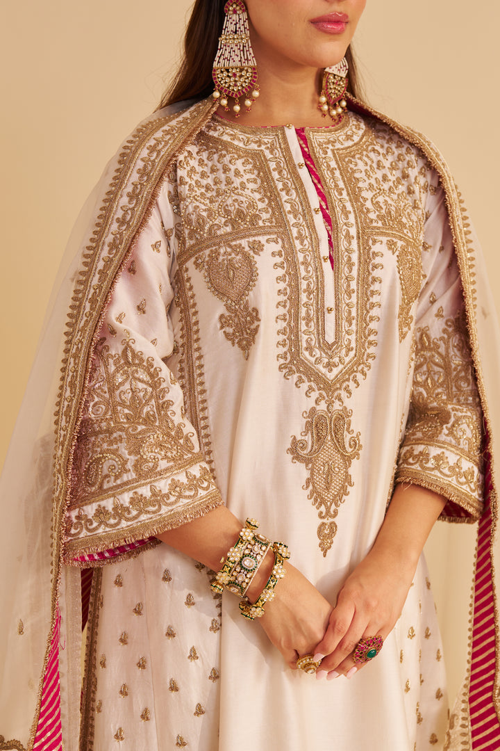 Woman wearing a cream-colored embroidered outfit with gold and red details on a beige background
