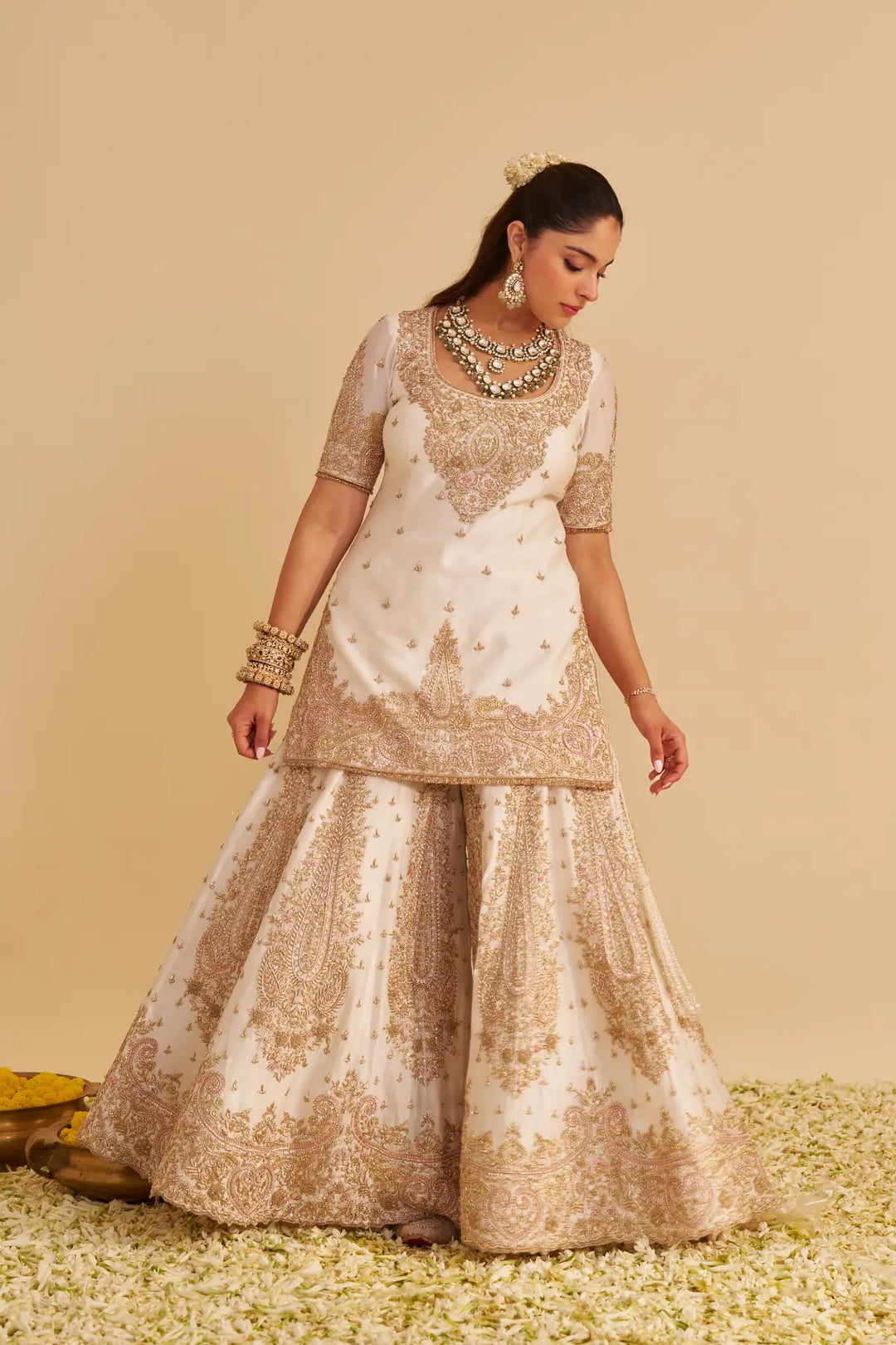 Woman wearing a traditional embroidered Sharara with gold and white patterns on a beige background