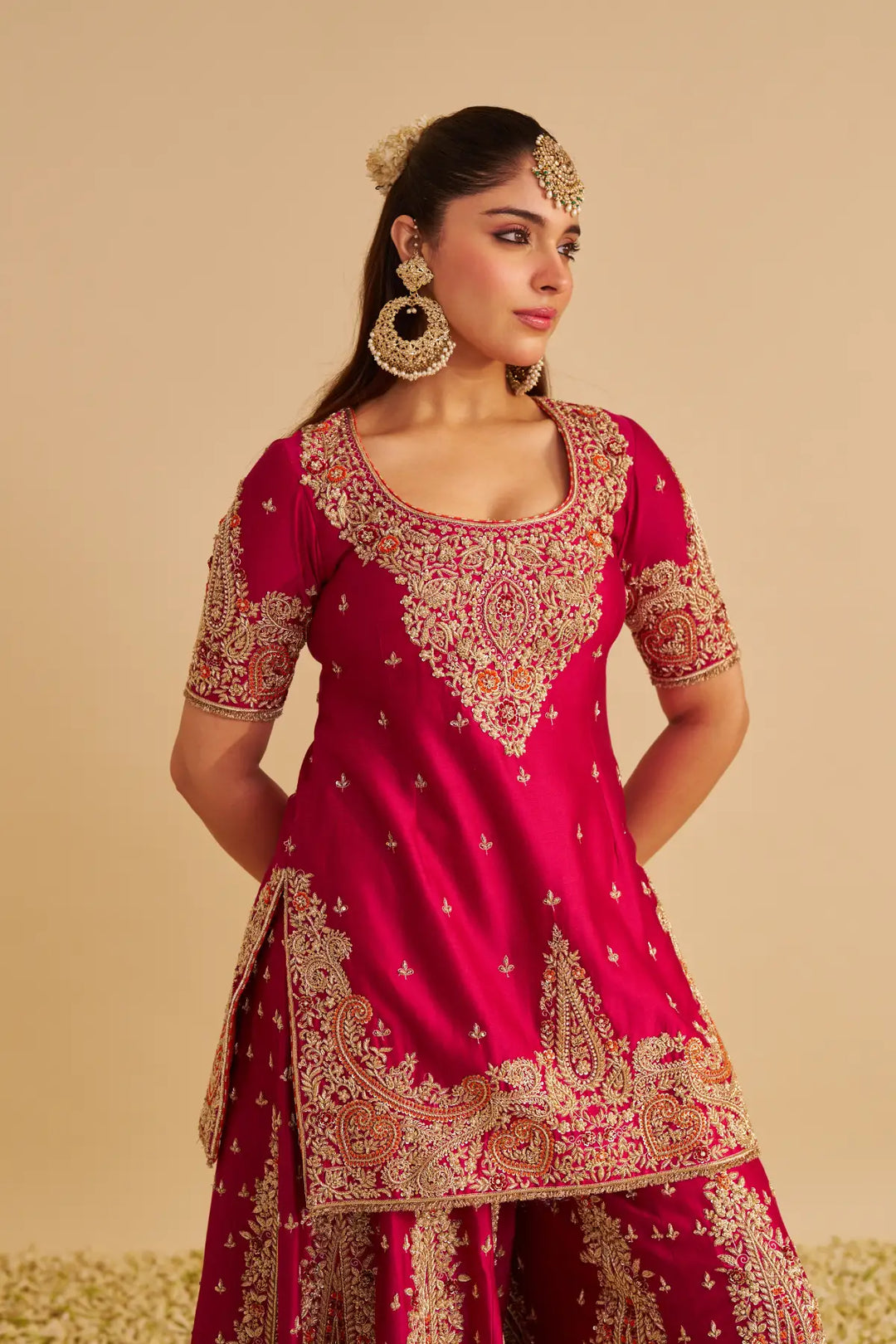 Woman wearing a traditional pink and gold embroidered Sharara on a beige background