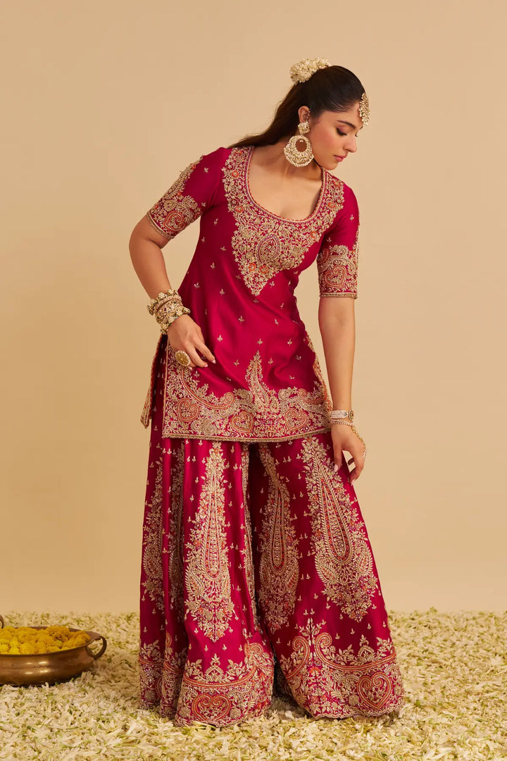 Woman wearing a red and gold embroidered traditional Sharara on a beige background