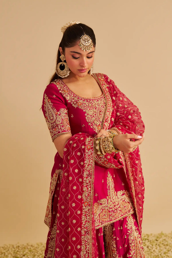 Woman in a red and gold traditional Sharara with intricate patterns on a beige background