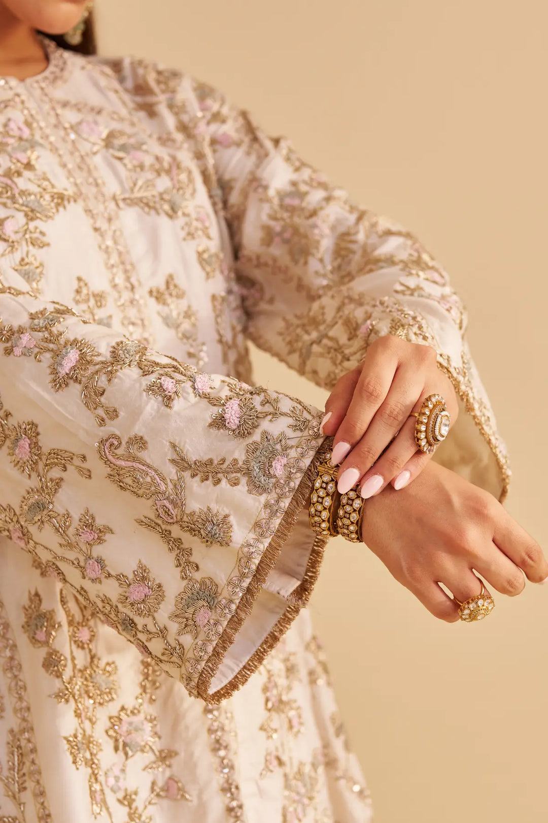 Close-up of a person wearing an intricately embroidered outfit with gold and beige colors on a beige background.