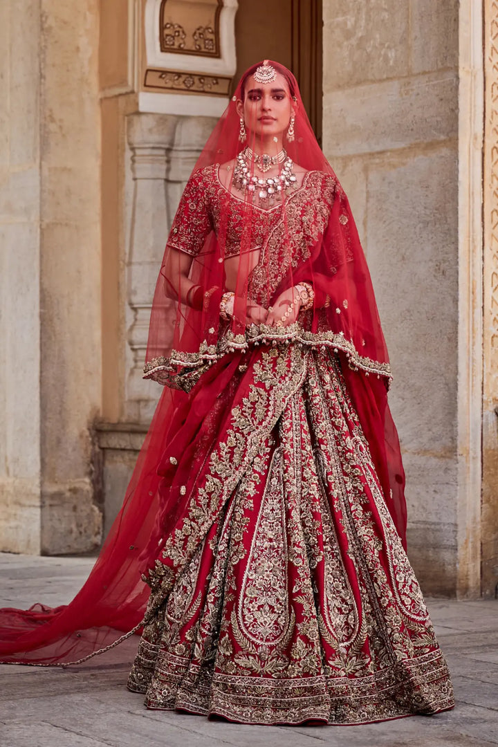 Woman in traditional red and gold bridal outfit with jewelry, standing against a stone wall.