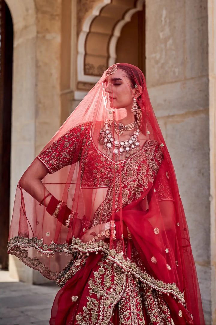 Woman in traditional red bridal outfit with jewelry, standing against a stone archway.