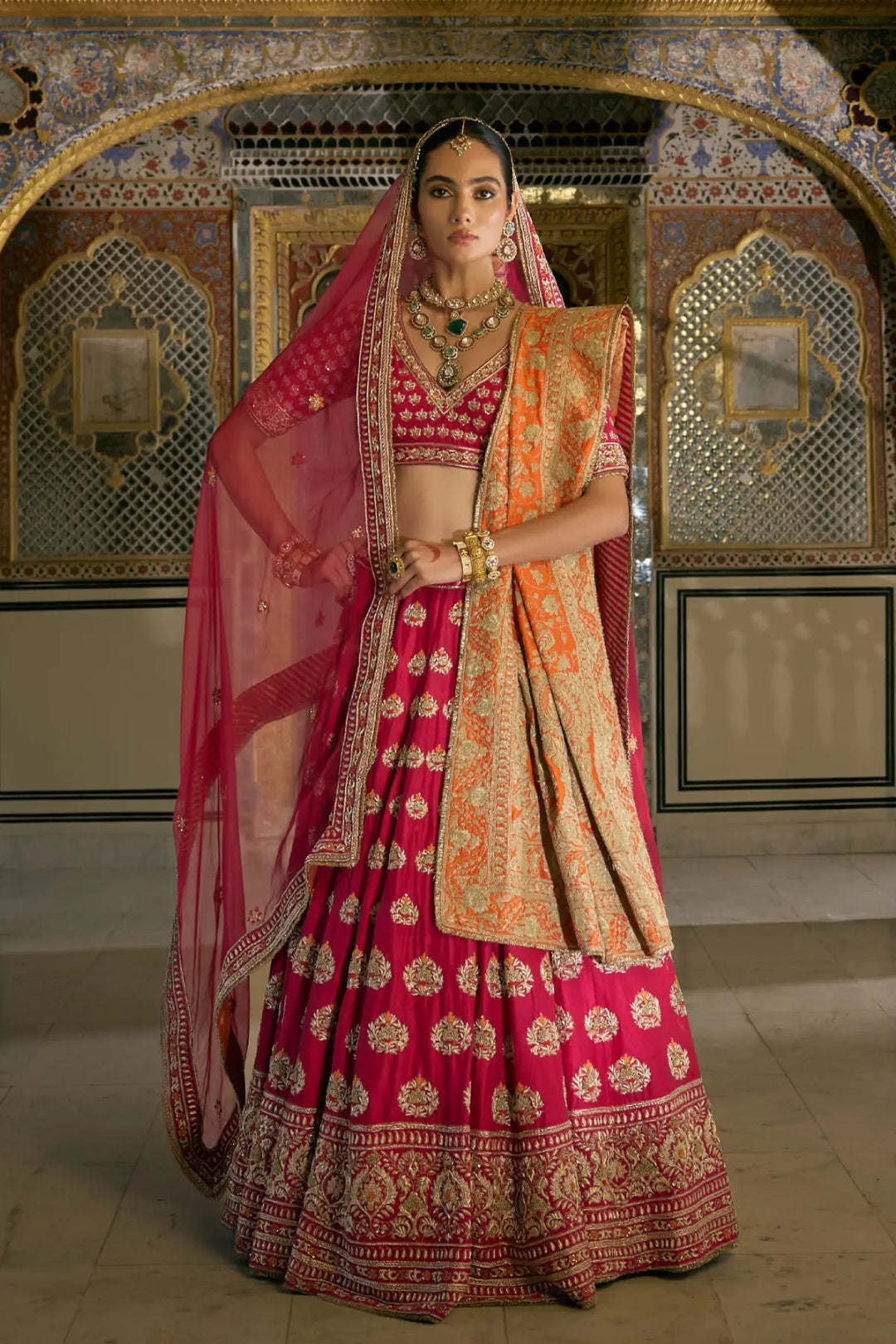 Woman in a traditional pink and gold embroidered lehenga with a matching dupatta, standing in an ornate room.