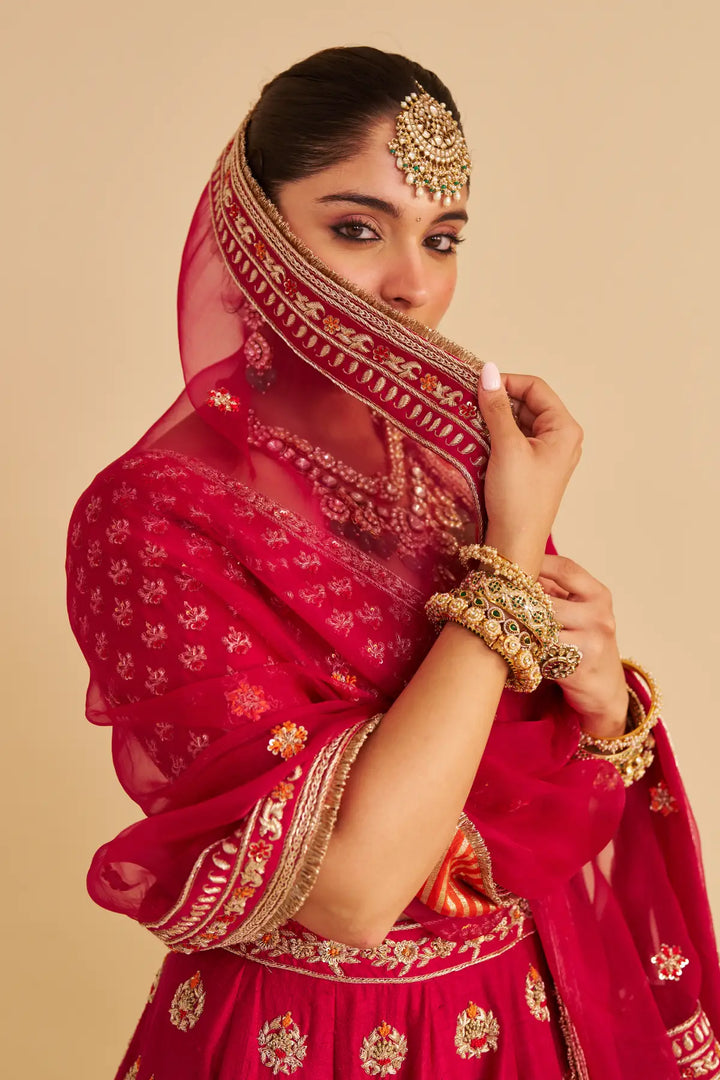 Woman in a red traditional outfit with gold embroidery and jewelry against a beige background