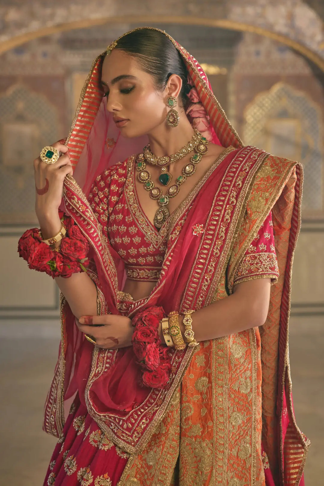 Woman in traditional red and gold embroidered outfit with jewelry, standing against a decorative background.
