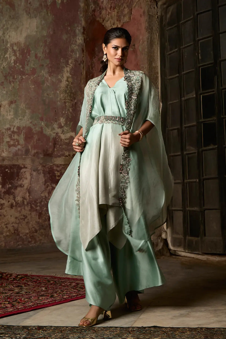 Woman in a light green traditional cape set standing against a textured wall.