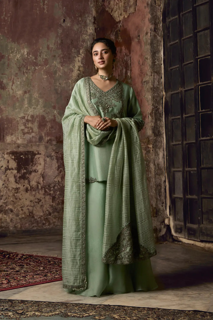 Woman in a green traditional sharara standing against a textured wall.