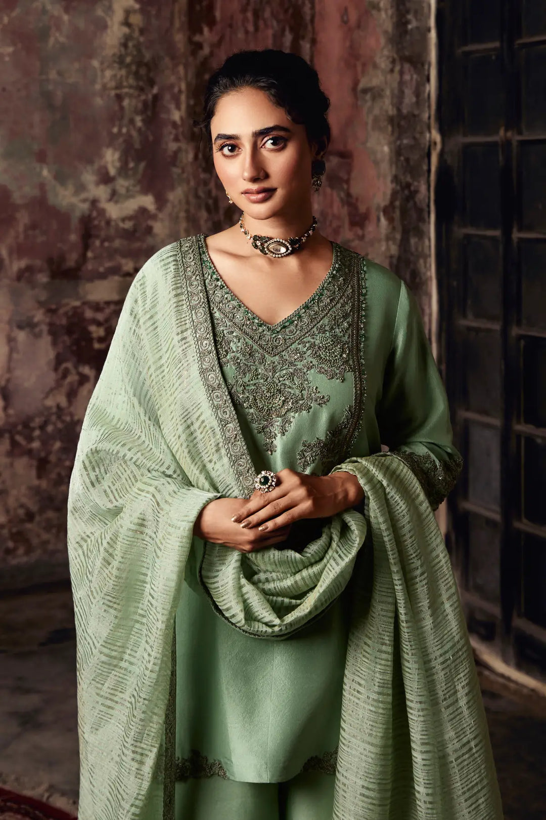 Woman in a green traditional sharara with a textured shawl against a rustic wall.