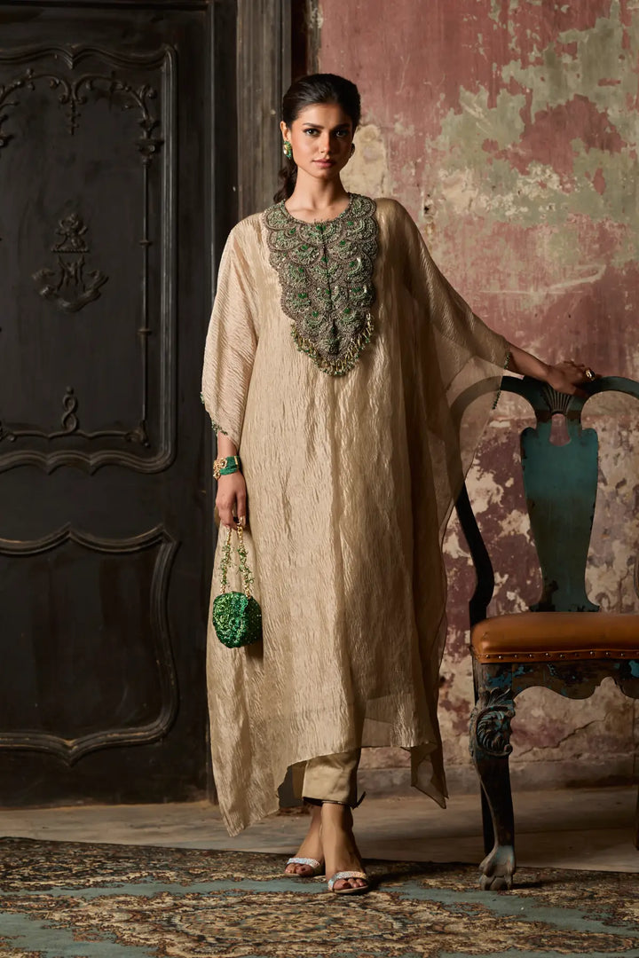 Woman in a beige traditional kaftan with intricate detailing standing in an ornate room.
