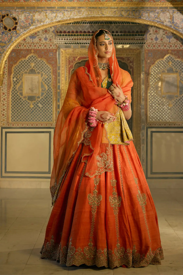 Woman in traditional orange and gold bridal lehenga with intricate patterns in a decorated interior setting.