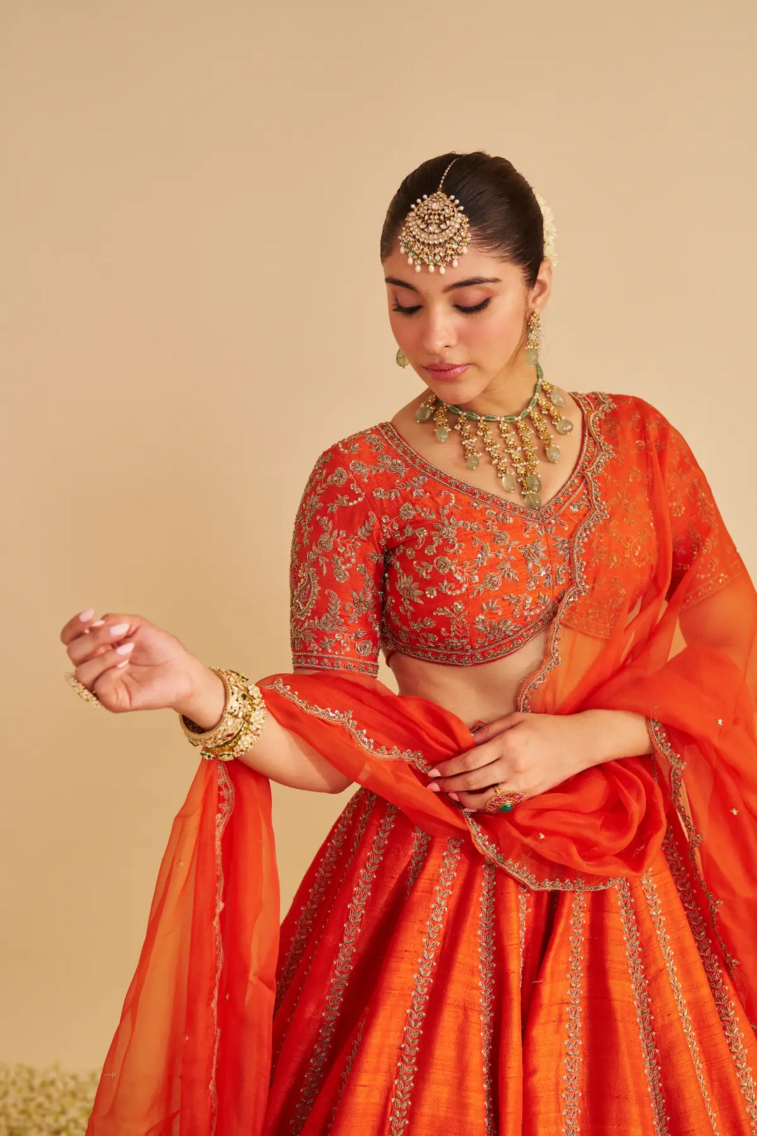 Woman in traditional orange embroidered lehenga with jewelry against a beige background