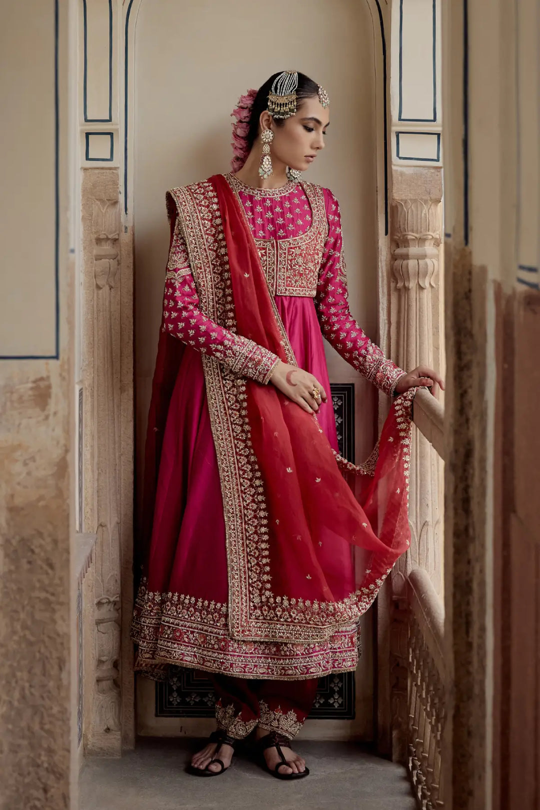 Woman in traditional pink and red embroidered Anarkali standing in a decorative doorway.