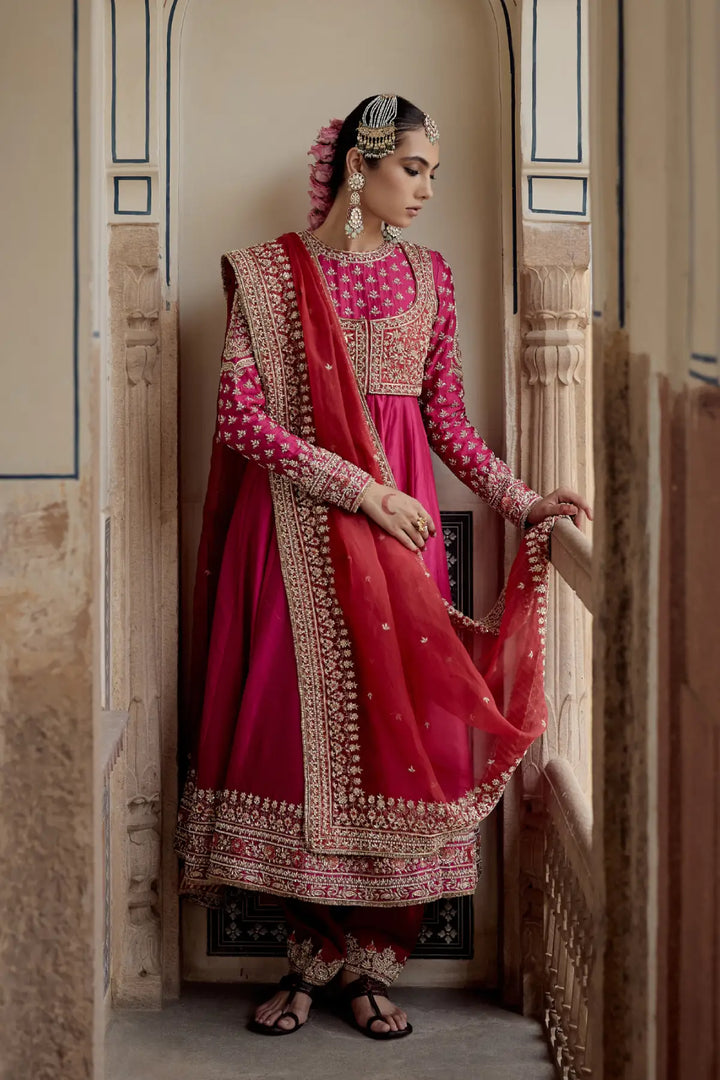 Woman in traditional pink and red embroidered Anarkali standing in a decorative doorway.