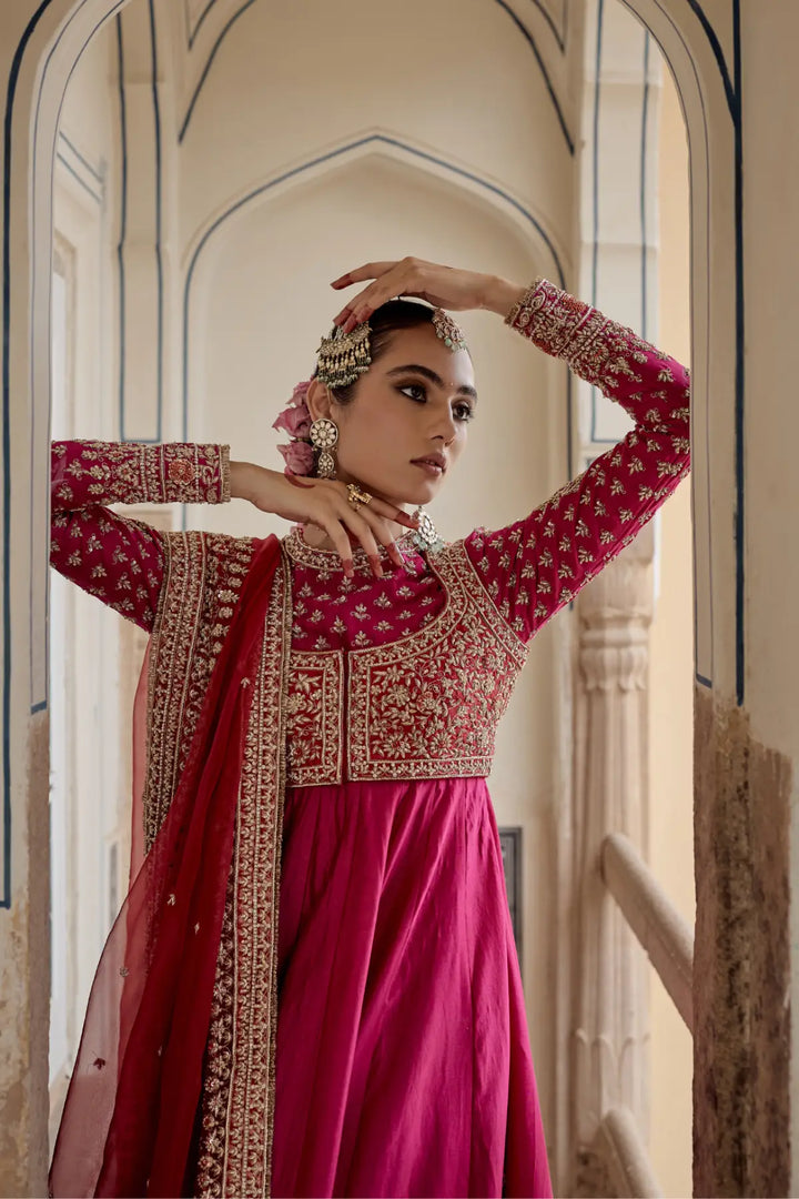 Woman in a traditional pink and red Anarkali with intricate patterns, standing in an archway.
