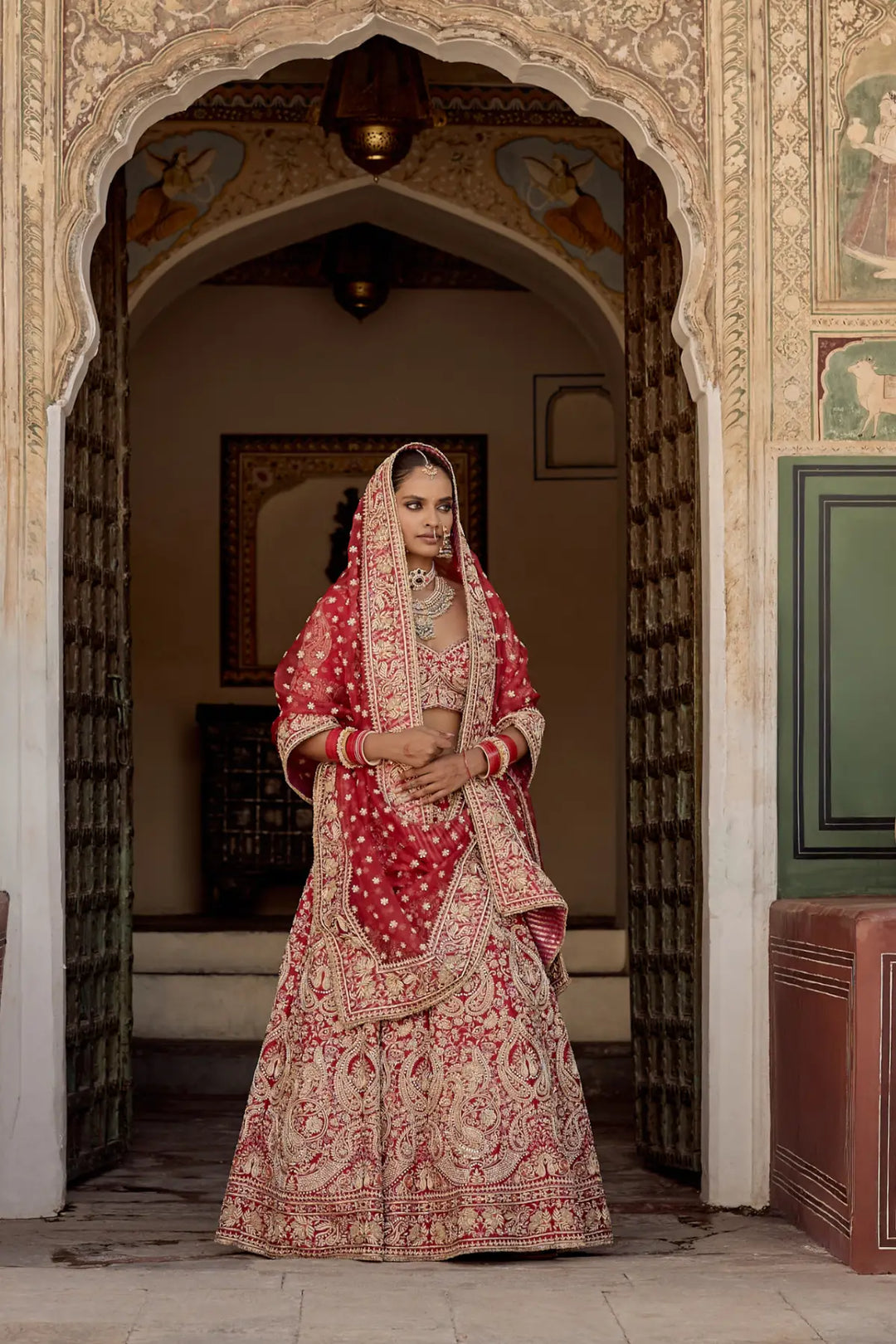 Woman in traditional red and gold bridal outfit standing in an ornate doorway.
