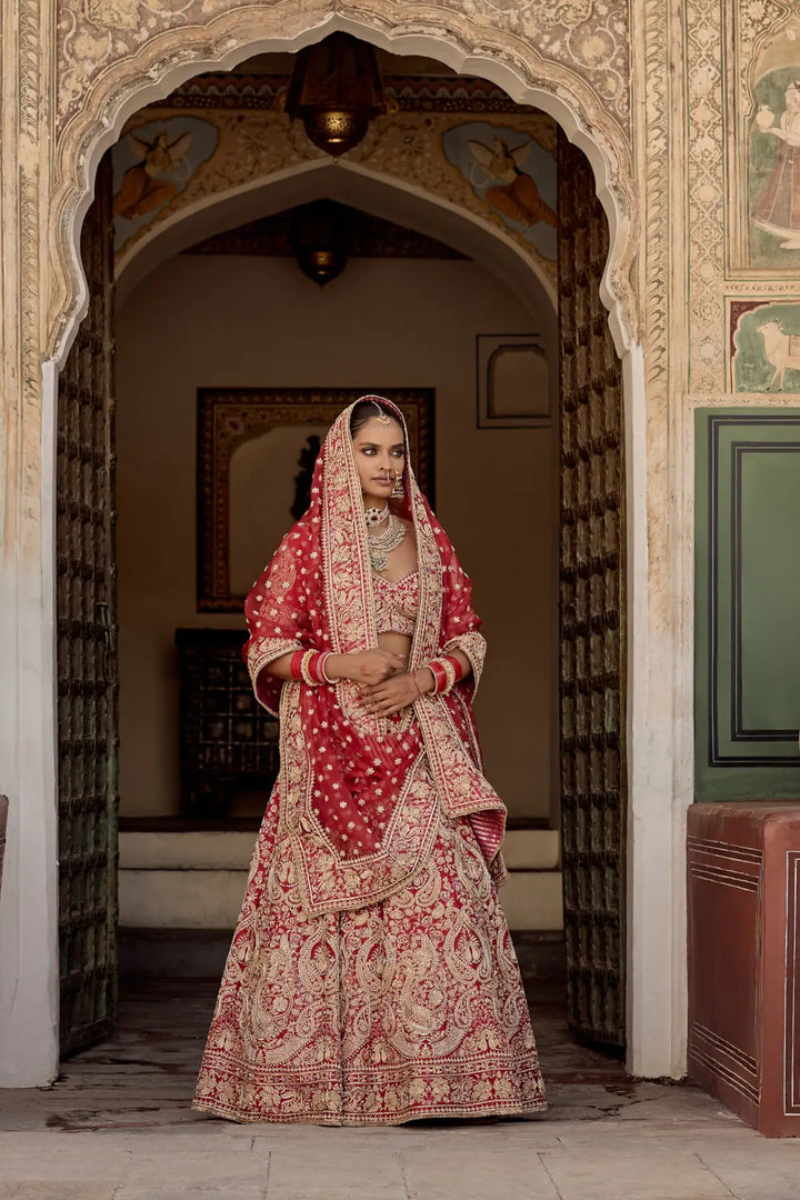 Woman in traditional red and gold bridal outfit standing in an ornate doorway.